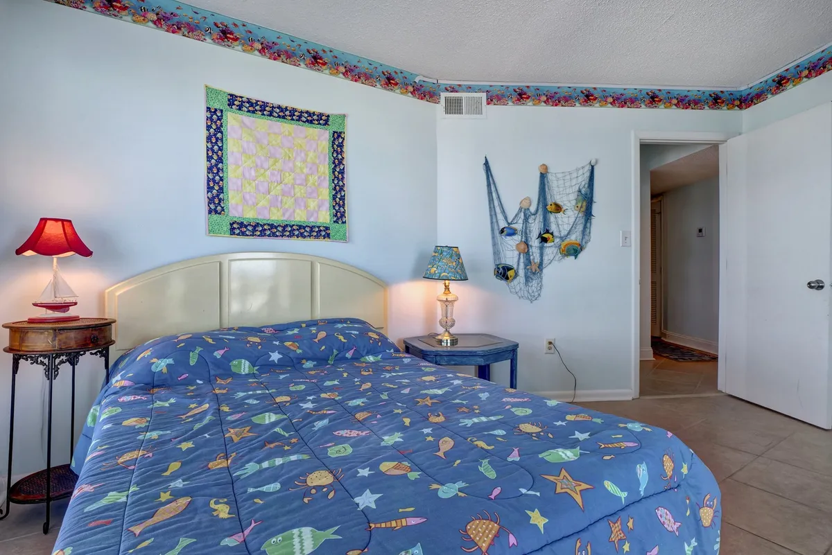 This image features a cozy bedroom with a colorful quilted bedspread, decorative wall hangings, a blue nightstand, and a warm lampshade. The cheerful and inviting atmosphere is accentuated by natural light filling the space, which is paired with beachy decor touches.