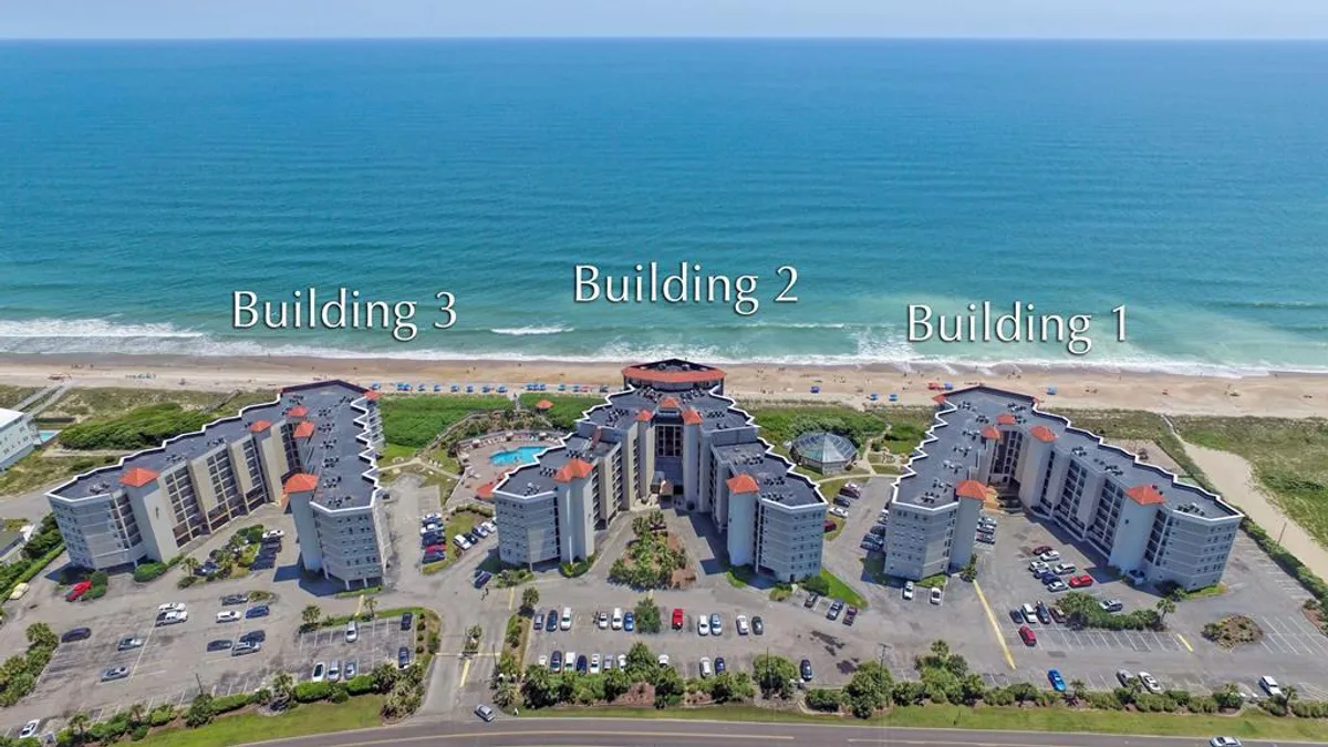 A wide exterior shot of the resort complex, featuring three distinct buildings, a sparkling swimming pool, and a sweeping ocean view in the background. The pool area is inviting, with lounge chairs and open space for groups to gather and relax.