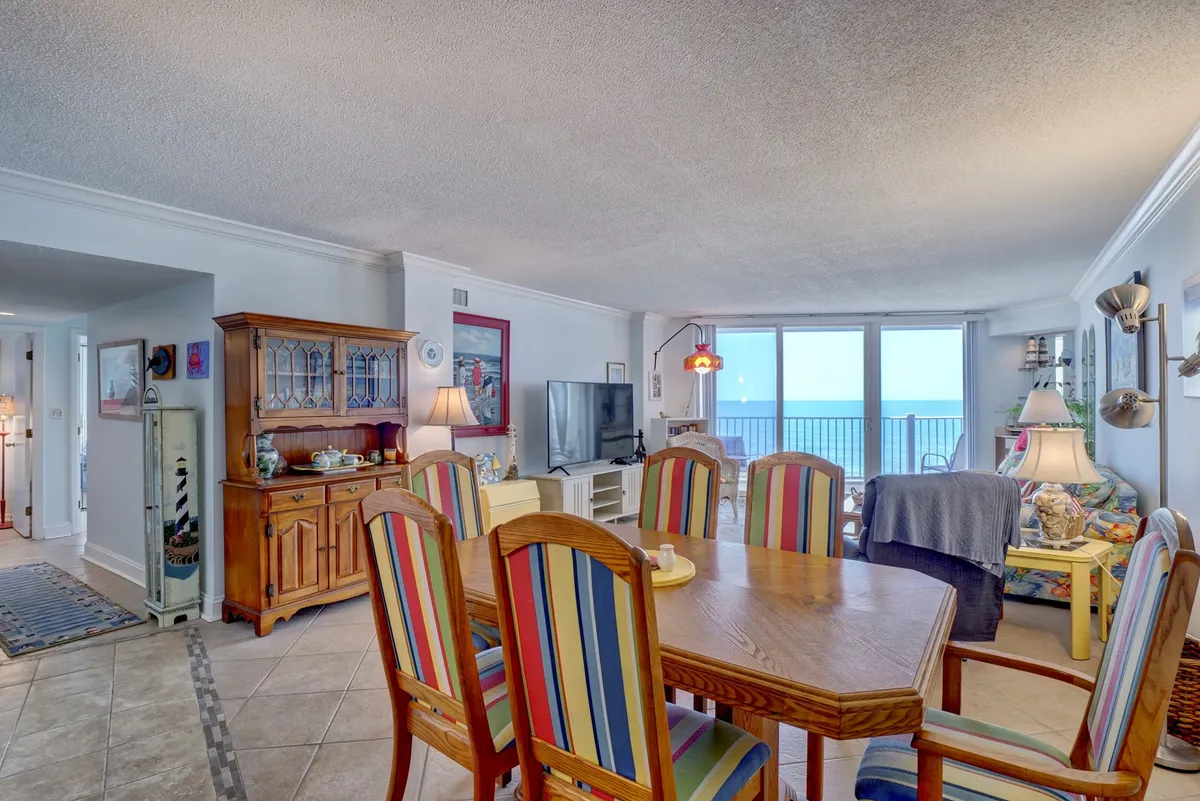 A relaxed dining area featuring a dining table with colorful chairs, large windows showcasing the ocean, and a wooden cabinet. The arrangement invites shared meals, board games, and social gatherings in a bright, cheerful setting.