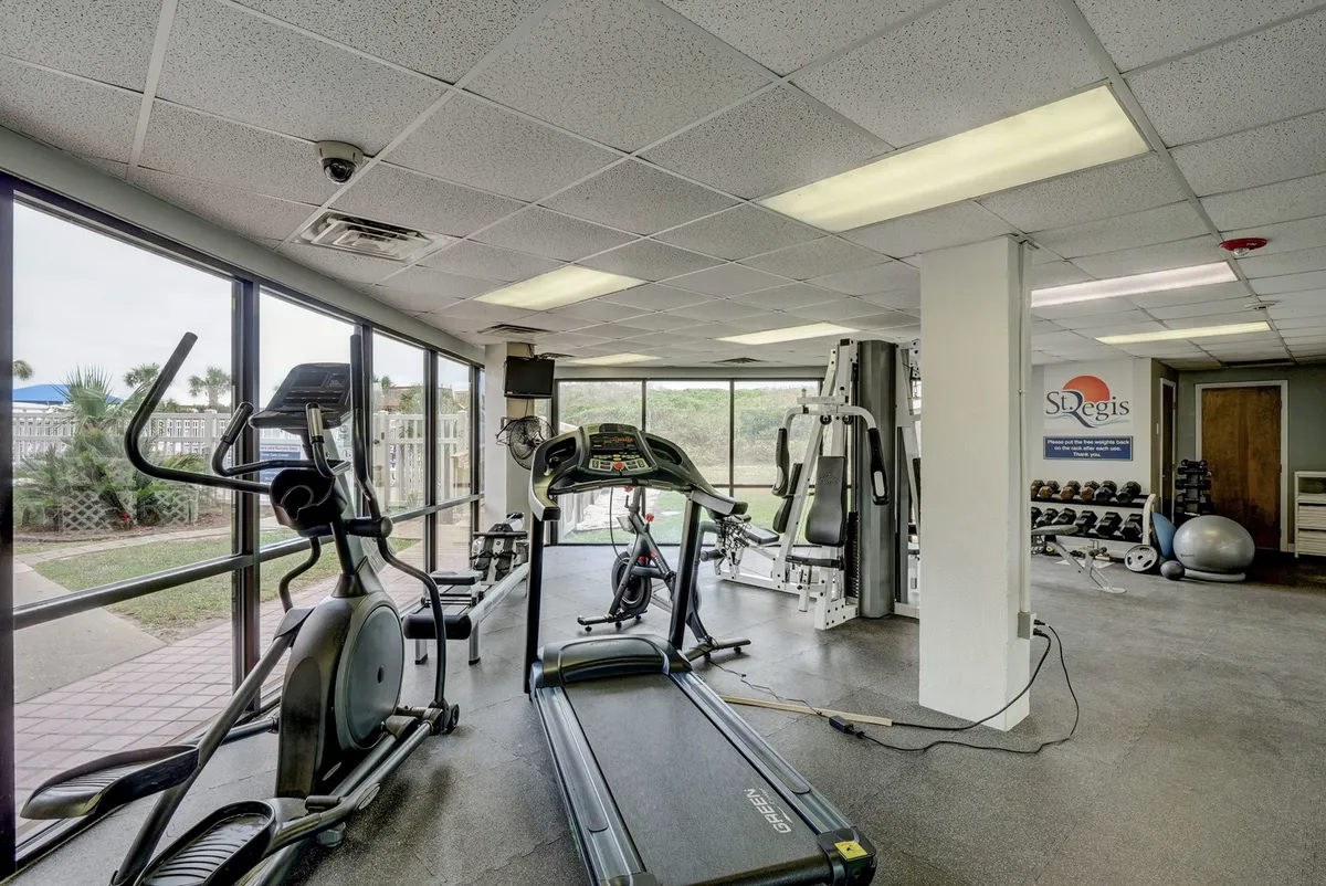 A bright, motivational fitness area equipped with a treadmill, elliptical machine, weight training equipment, and large windows that flood the room with natural light. The space is designed for both individual workouts and group fitness activities.