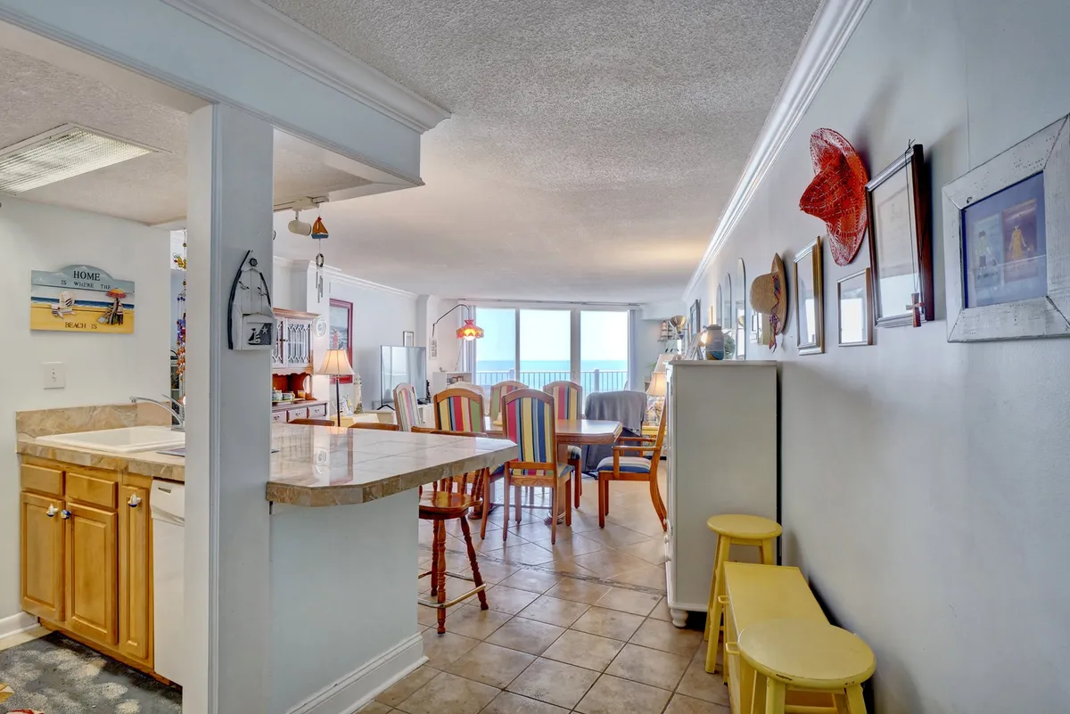 This image captures a bright, open-plan dining and living area featuring colorful dining chairs, large windows with a sweeping ocean view, an open kitchen, and lively wall art. Sunlight pours in, highlighting the communal table set for family meals and gatherings, while the arrangement encourages conversation and togetherness.