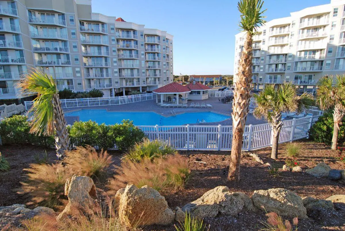 This image features a sparkling swimming pool surrounded by lush greenery and palm trees, with vacation-style architecture visible in the background. The tranquil environment is perfect for relaxation, sunbathing, and socializing, evoking a classic resort atmosphere.
