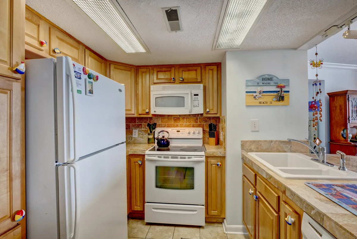 A cheerful kitchen with wooden cabinets, white appliances, a kitchen sink, and a decorative wall sign. The space is well-lit and clearly set up for meal prep and casual dining, suggesting a welcoming environment for shared cooking and family gatherings.