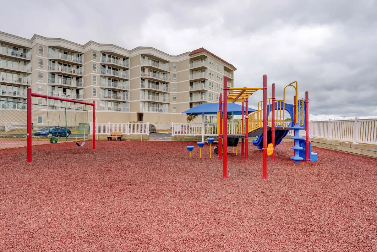 A lively outdoor playground surrounded by a residential building, showing children playing, families gathering, and the joyful atmosphere of community recreation. The scene is vibrant and welcoming, reflecting the property’s family-friendly amenities.