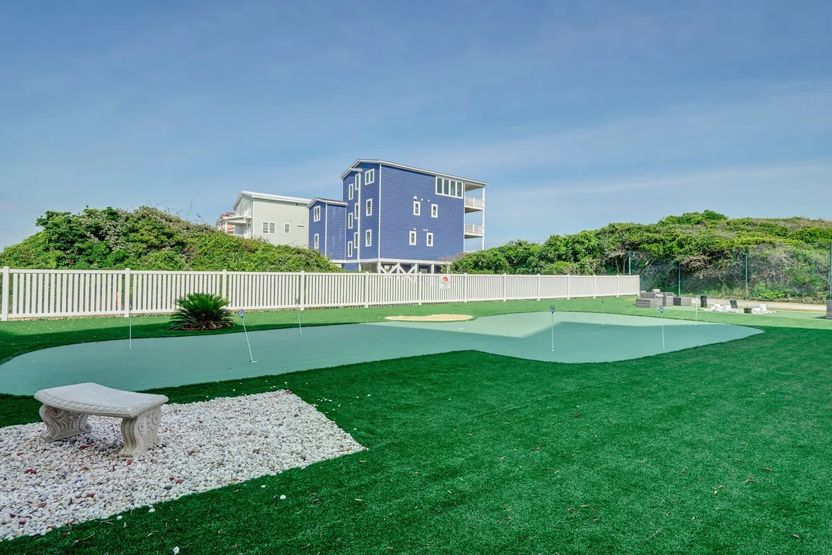 An exterior view of a well-maintained putting green surrounded by lush landscaping and a blue house in the background. The area invites relaxation and casual recreation, perfect for golf practice or family fun.