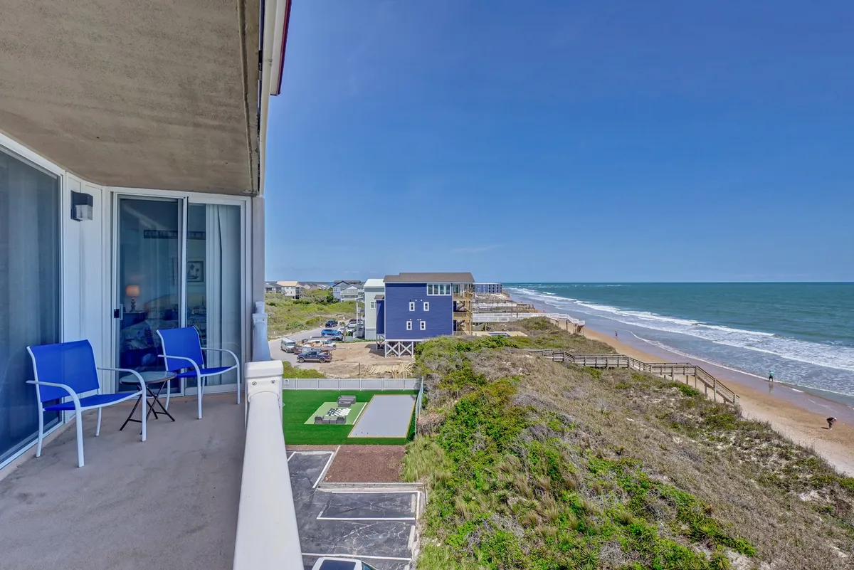 This image showcases a serene oceanfront balcony with comfortable seating, overlooking the nearby beach and a green lawn. The vantage point provides a clear view of the coastline, inviting guests to enjoy sunrises, ocean breezes, and the relaxing ambiance of coastal living.