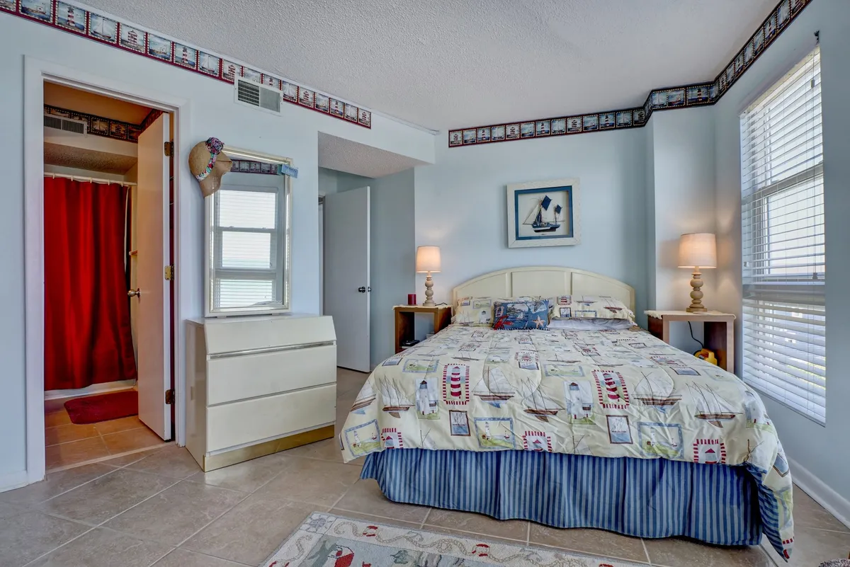 A bedroom with nautical-themed bedding, wooden side tables topped with lamps, framed nautical artwork on the wall, and a glimpse into an adjoining bathroom with a red shower curtain. The room is bright with natural sunlight, combining comfort and maritime style.