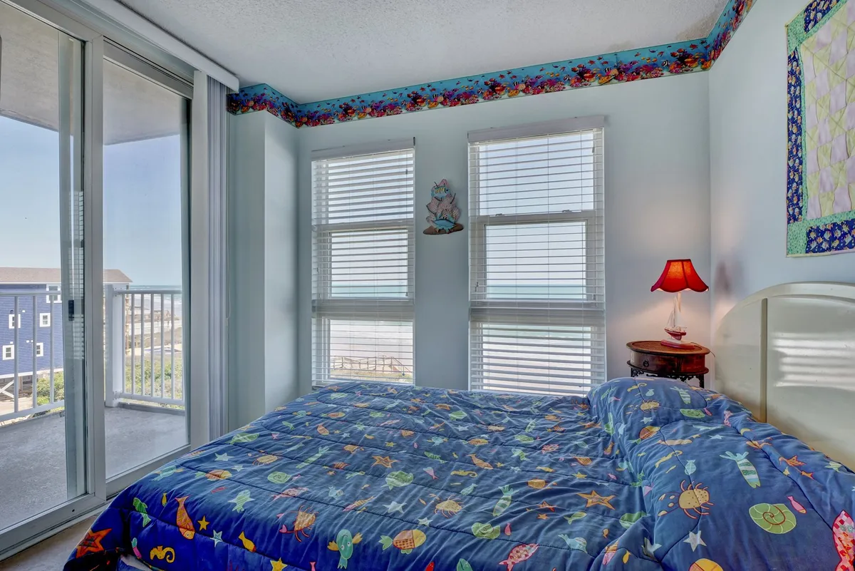 A bright, playful bedroom with colorful bedding, large windows allowing in ample sunlight, a decorative lamp, and clear ocean views. The room feels open and airy, enhancing its relaxing, coastal charm.