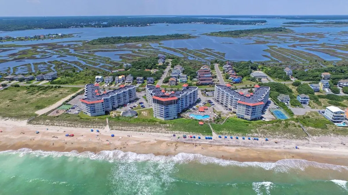 A wide view of a beachfront property with a swimming pool at the foreground, colorful beach umbrellas, ocean waves, and surrounding marshlands. The scene is lively and inviting, suggesting accessibility to both the beach and pool facilities, perfect for family gatherings and seaside fun.