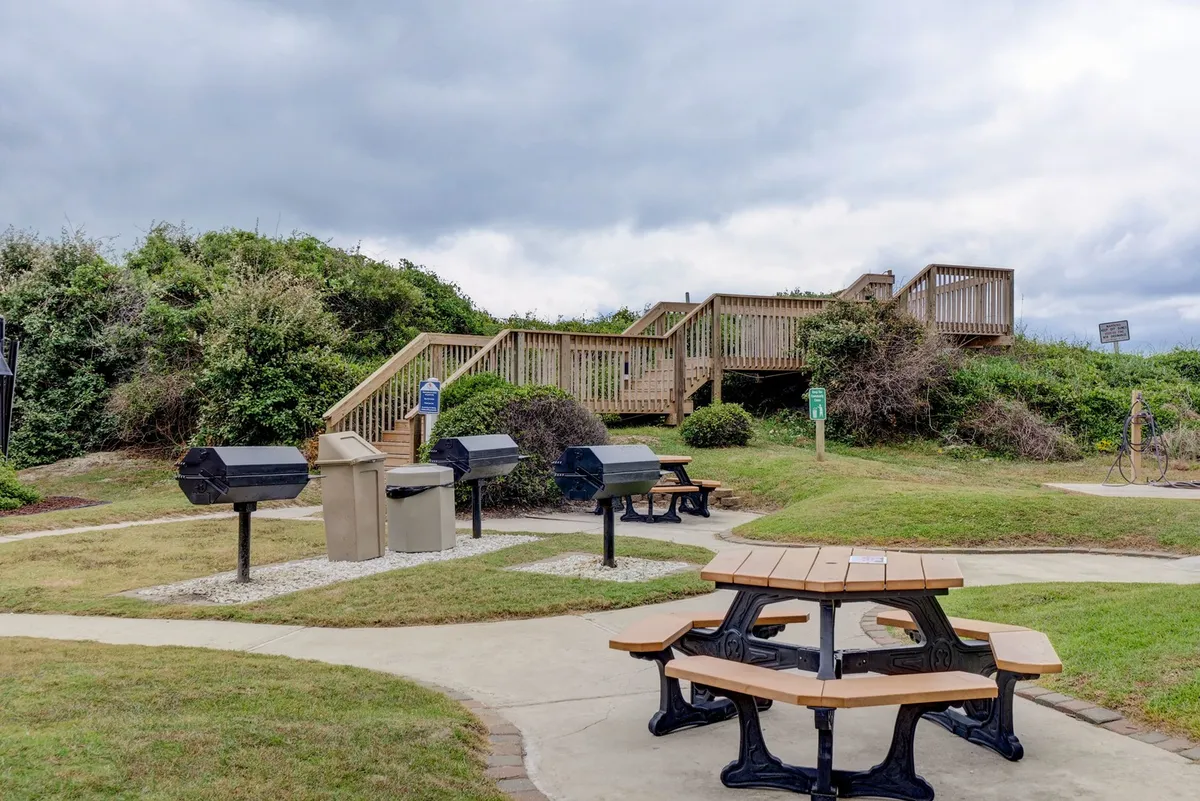 A relaxed outdoor communal area with picnic tables and grilling stations surrounded by greenery. The space is set up for barbecuing, picnicking, and group gatherings, encouraging social interaction and outdoor dining.