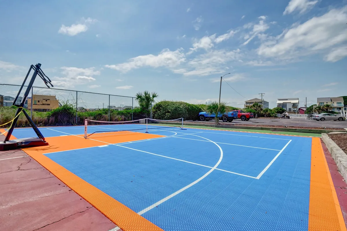 An outdoor recreational area with a basketball hoop and tennis net set on a vibrant, colorful court surface. The court is surrounded by open sky and ample space for group sports and activities.