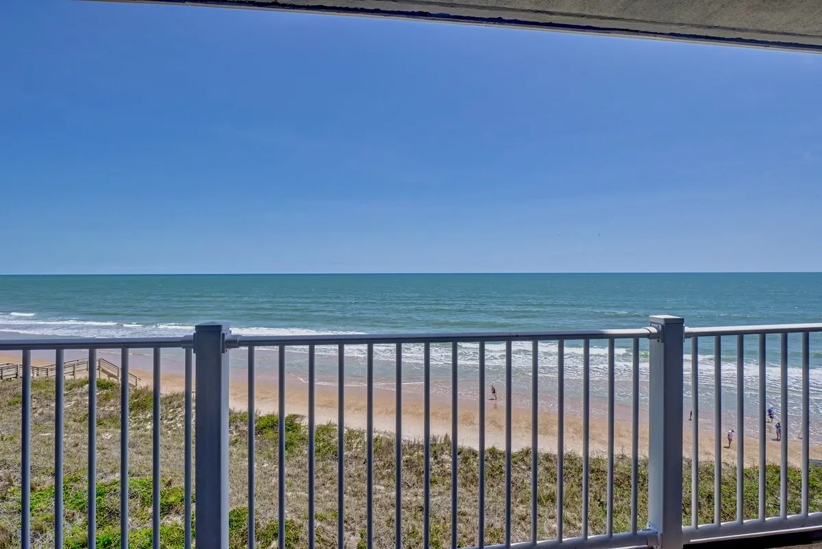 The image showcases a stunning oceanfront view from the condo's private balcony, with the sweeping expanse of North Topsail Beach and the rolling surf visible below. The balcony railing frames the tranquil seascape, inviting guests to relax and soak up the sun or greet the sunrise. The scene exudes calm and endless vacation potential, with the sandy beach and gentle waves setting a perfect backdrop for relaxation and beachside adventures.
