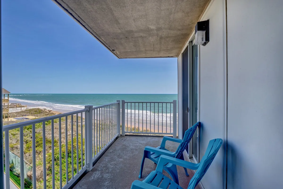 A serene outdoor balcony with blue chairs overlooking a stunning beach view and protective railing. The tranquil setting is ideal for soaking in ocean breezes, enjoying sunsets, or meditating to the sound of the waves.
