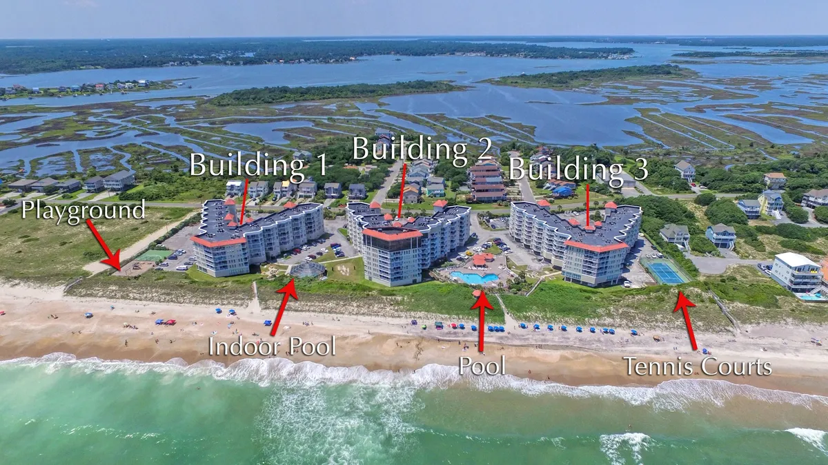 A panoramic exterior view of the resort complex, showcasing multiple buildings, an indoor and outdoor pool, tennis courts, and a playground set against the backdrop of the ocean. Children can be seen playing, and the pools and recreation areas are visible.